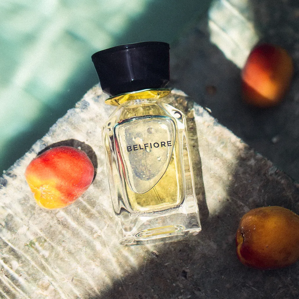 Parfümflakon BELFIORE liegt mit Pfirsichen nahe Wasser.